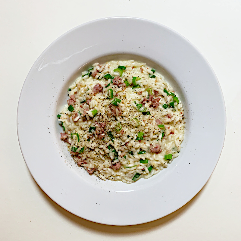 Ein weißer Telle auf weißer Tischdecke mit Salsiccia-Risotto mit grünen Frühlingszwiebeln