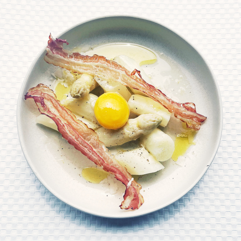 Ein Teller mit weißem Spargel, Speck, Ei und Parmesan angerichtet.